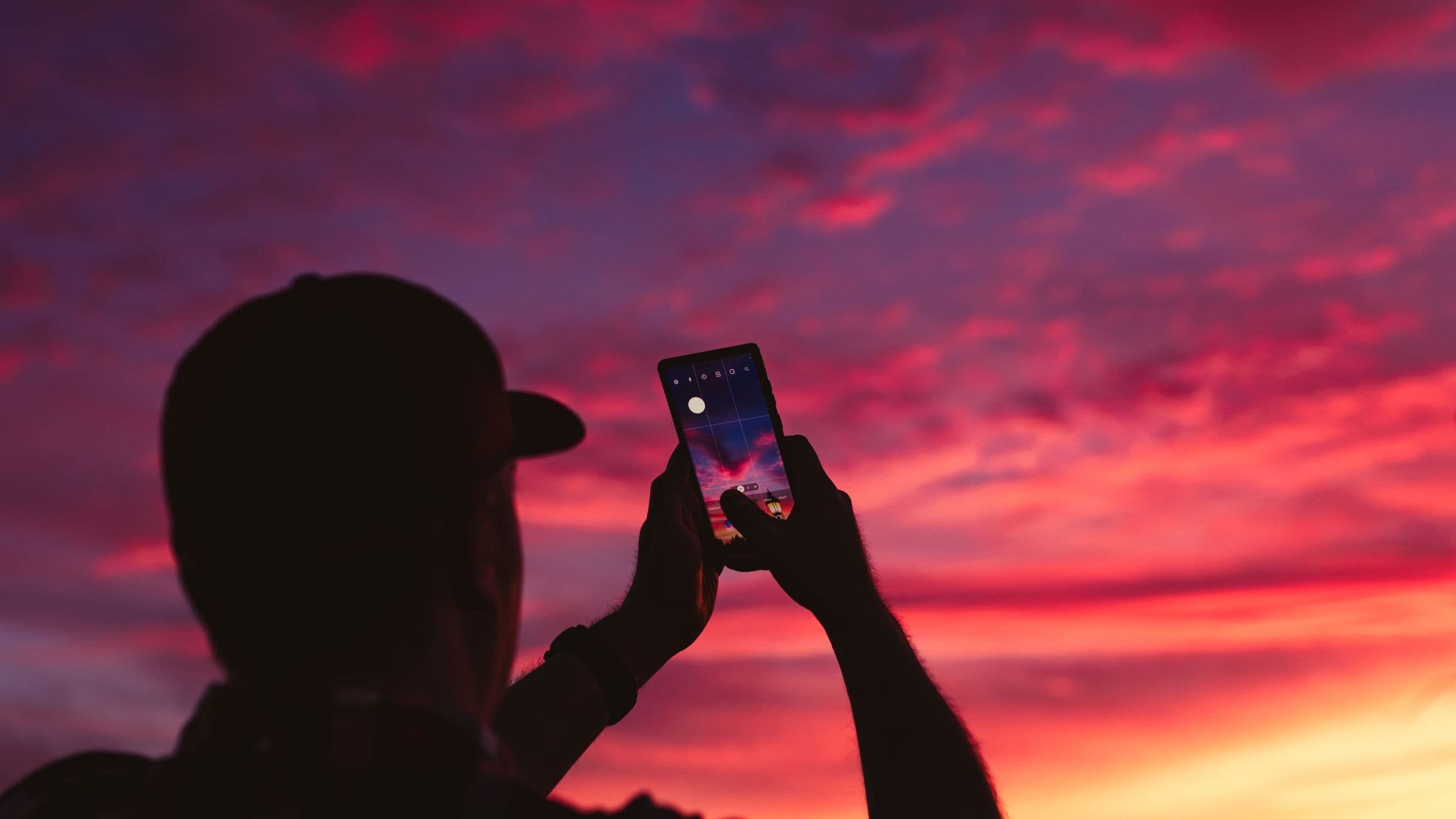 The silhouette of a person holding up a smartphone against a vivid magenta and orange sunset sky.