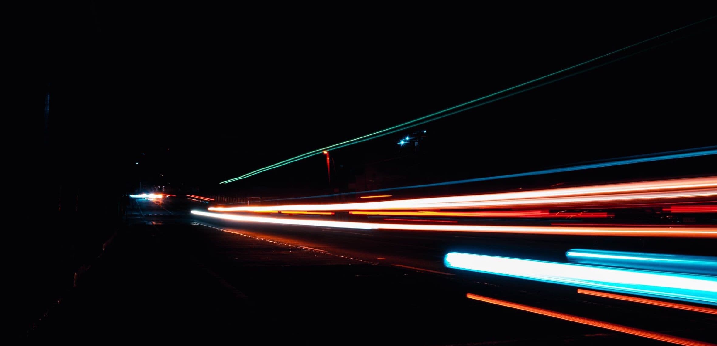 Long exposure light trails streaking along a dark road at night, in orange, white and blue.